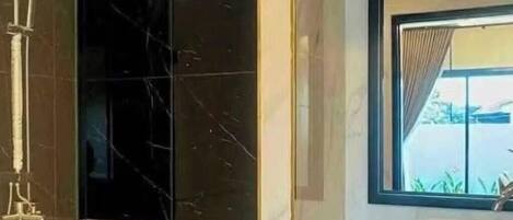 Un baño con pared de mármol negro, cabina de ducha de vidrio, espejo con marco dorado y una ventana con cortinas.