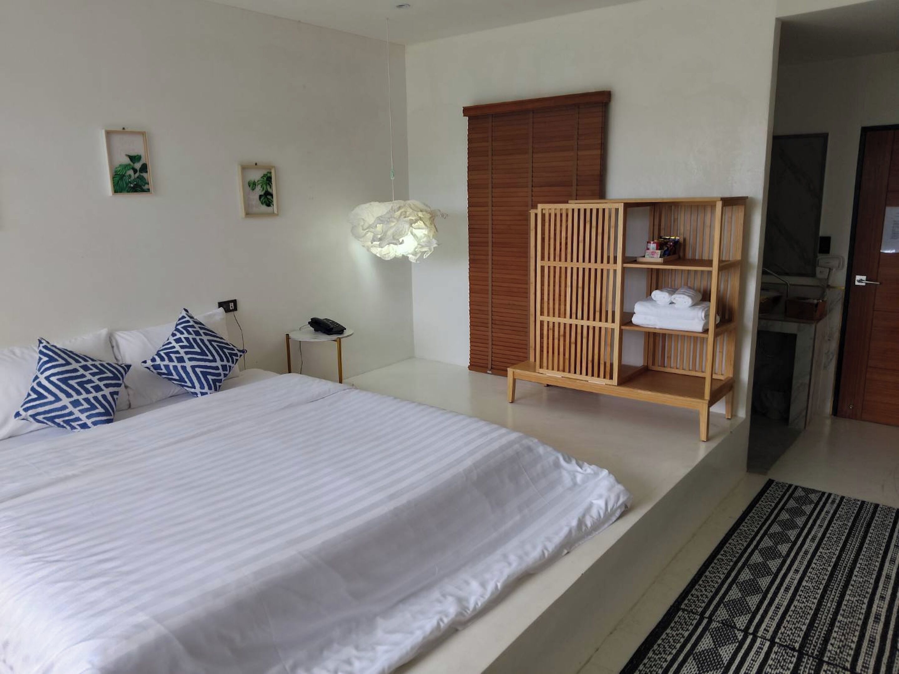 Una habitación de hotel con una cama grande, un armario de madera, una mesita de noche con lámpara y un estante pequeño con toallas.