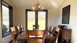 A dining room with a large wooden table and chairs, a chandelier, and a view of a yard through large windows.