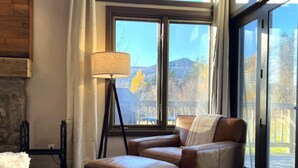 A cozy living room with a leather sofa, a floor lamp, and a view of a deck and mountains through large windows.