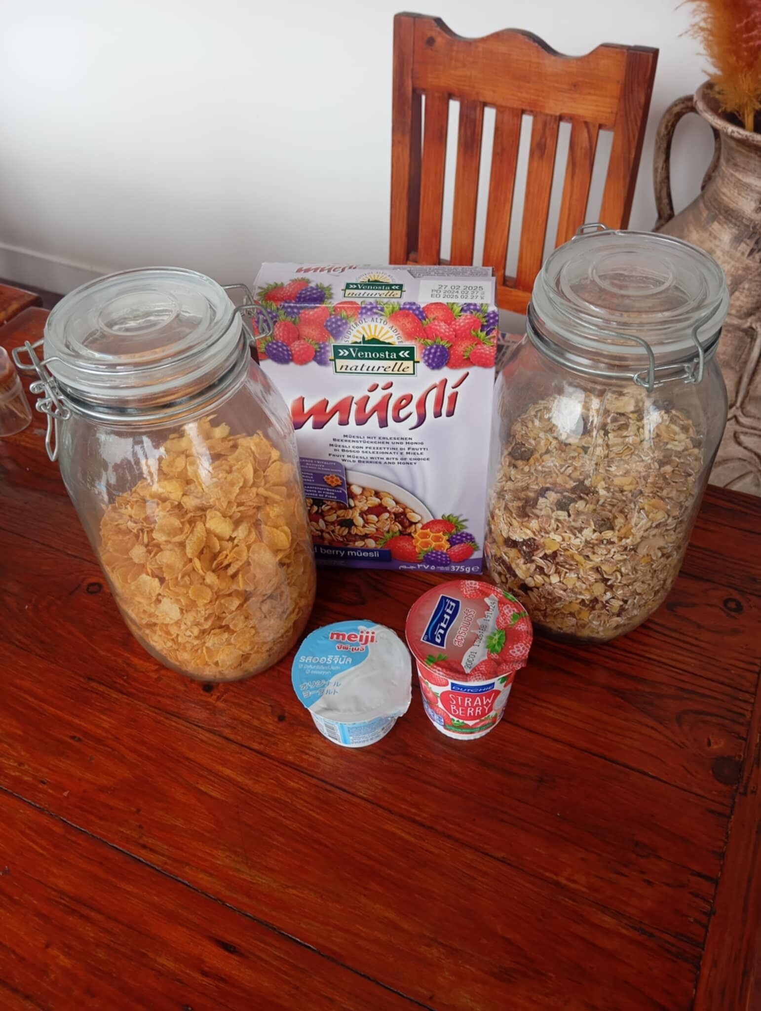 Een houten tafel met twee glazen potten gevuld met ontbijtgranen, een pak muesli en twee kleine bakjes yoghurt.