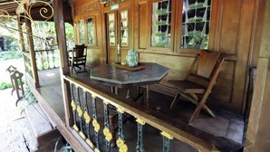 Une véranda en bois avec une table et des chaises, une balustrade décorative et une fenêtre donnant sur un paysage verdoyant.