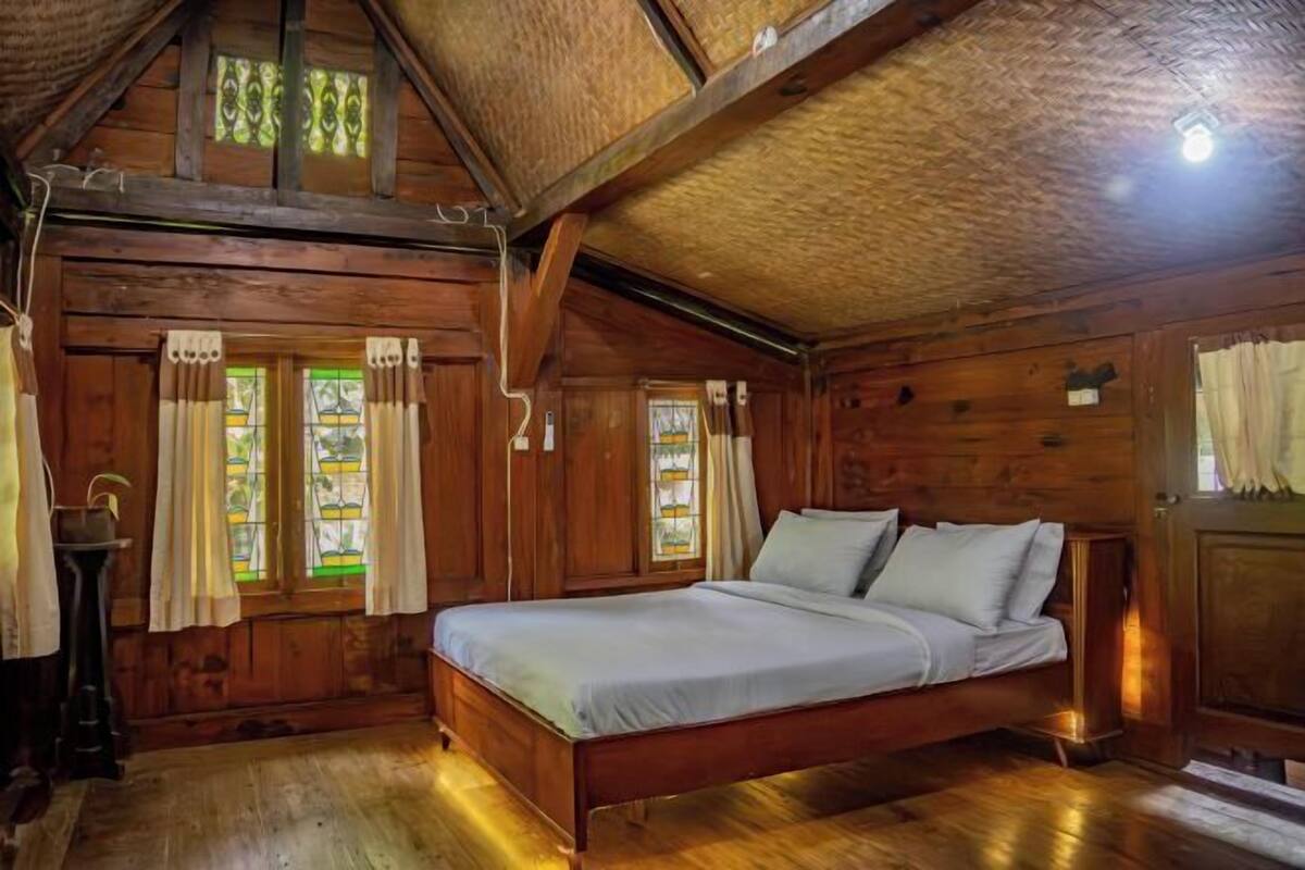 A wooden cabin room with a bed, windows with curtains, and a wooden ceiling.