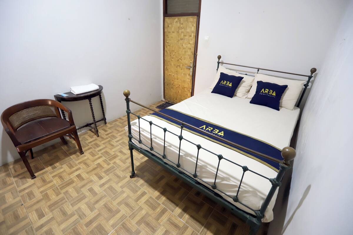 A bed with a metal frame and a mattress labeled 'AR3A'. There is a wooden chair and a small table with a white object on it.