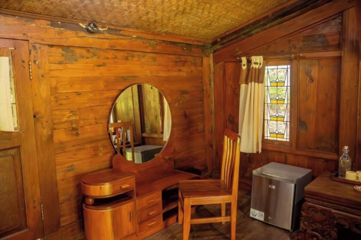 A wooden room with a round mirror, a wooden chair, a small refrigerator, and a window with curtains.