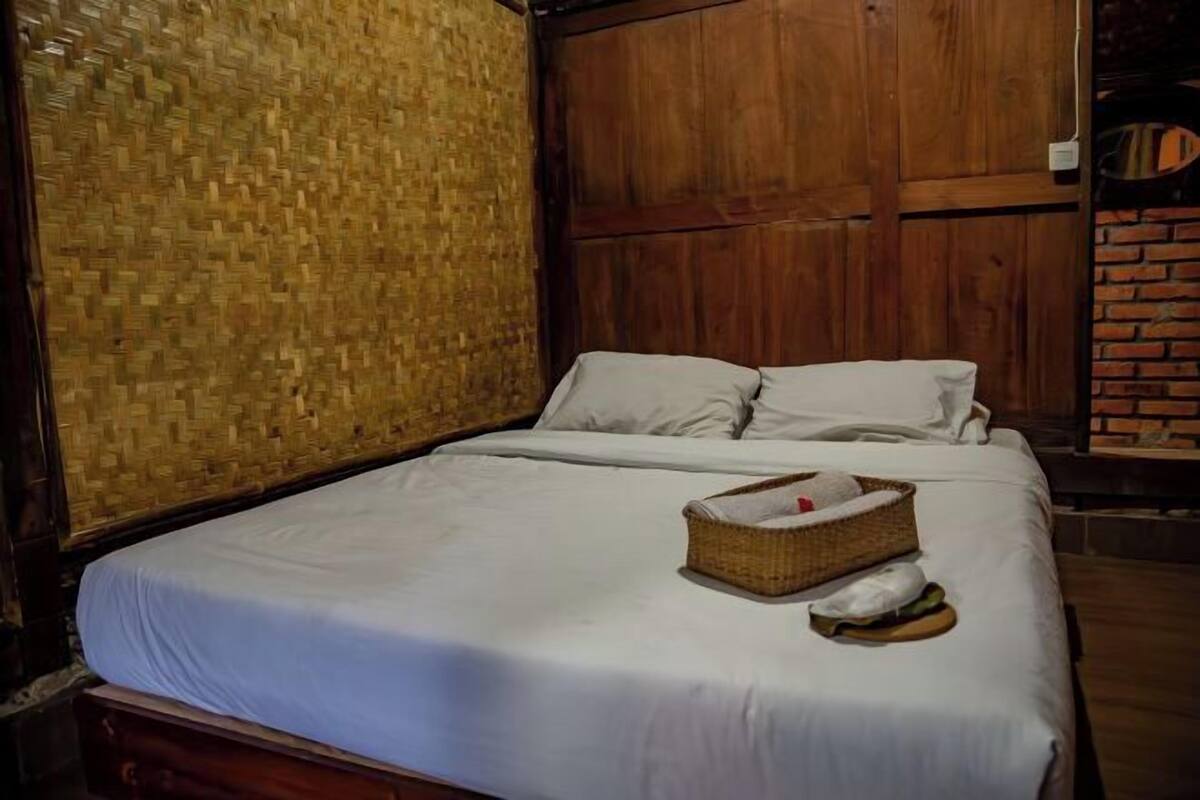 A single bed with white linens, a wicker basket with a towel, and a brick wall.