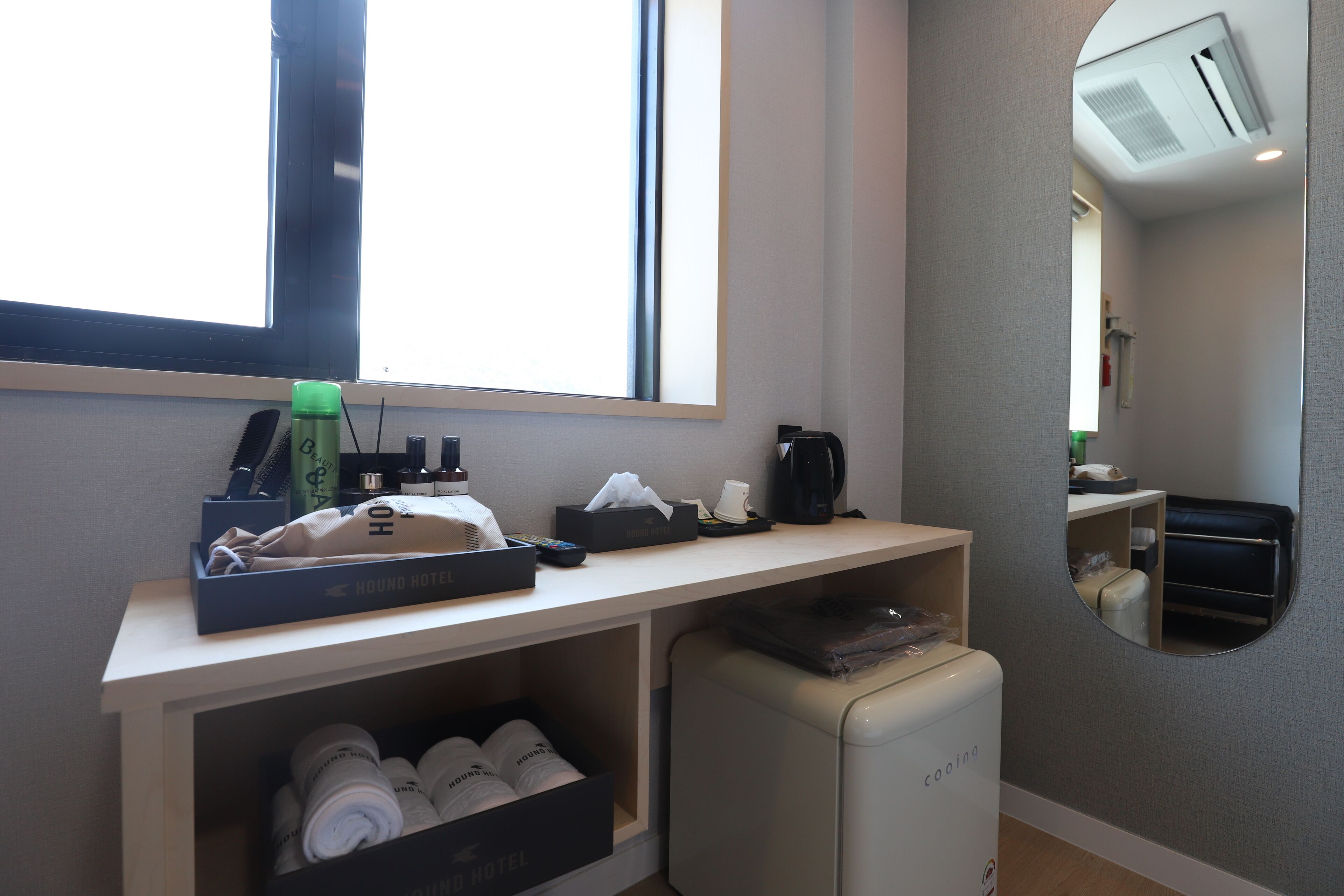 A hotel room with a desk, a mirror, a small fridge, and a window.
