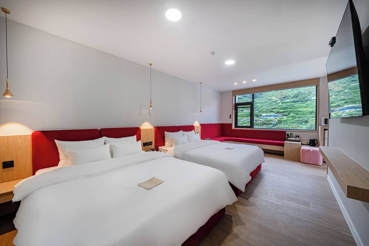 A hotel room with two beds, a red accent wall, a large window with a view of greenery, and a flat-screen TV mounted on the wall.