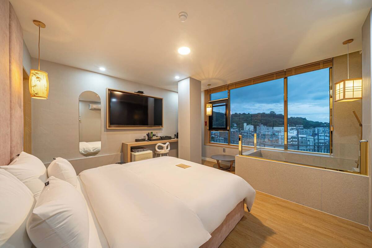 A modern hotel room with a large bed, a desk, a mirror, and a large window offering a city view.