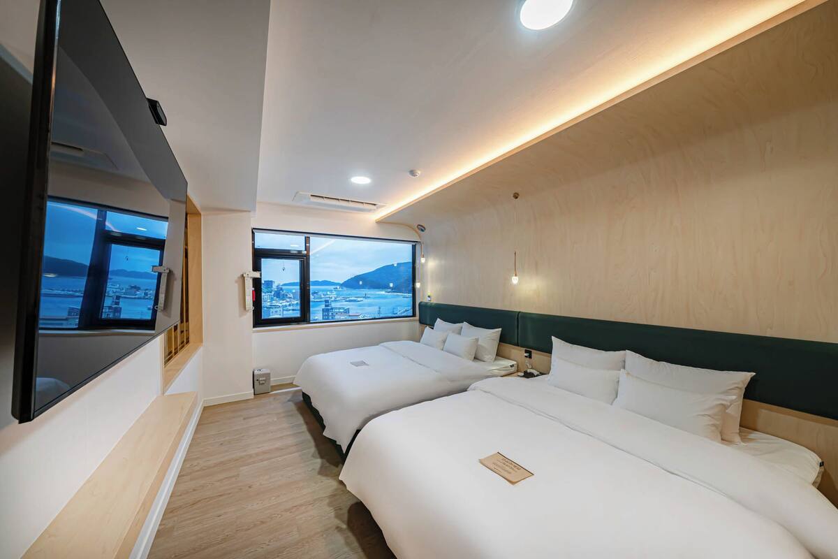 A hotel room with two beds, a large window with a city view, and a flat-screen TV mounted on the wall.