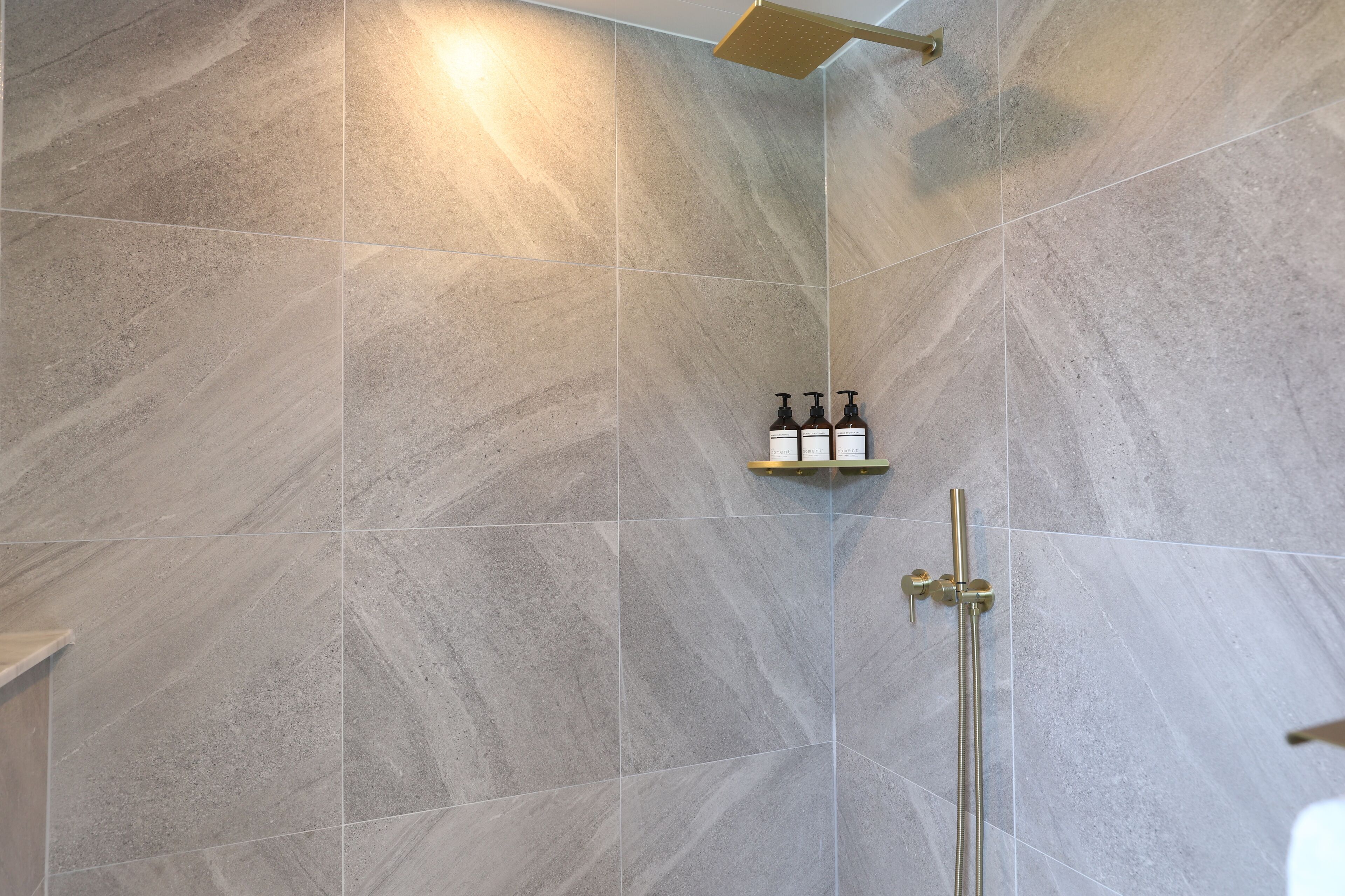 A shower area with grey tiles, a gold showerhead, and a shelf with three bottles.