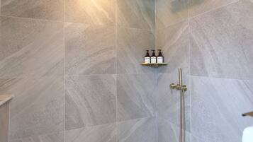 A shower area with grey tiles, a gold showerhead, and a shelf with three bottles.