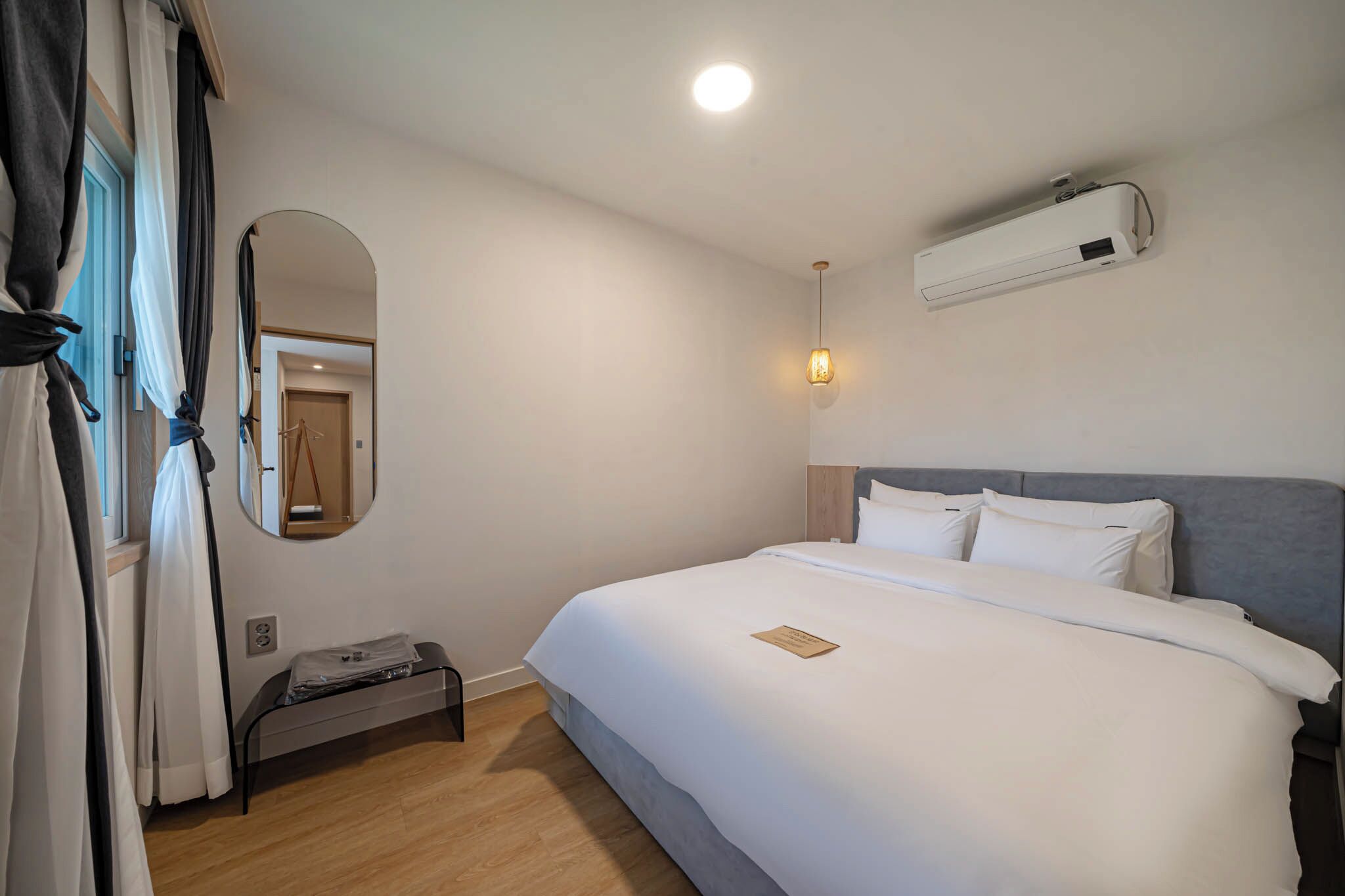 A hotel room with a bed, a bedside table, a mirror, and an air conditioning unit.