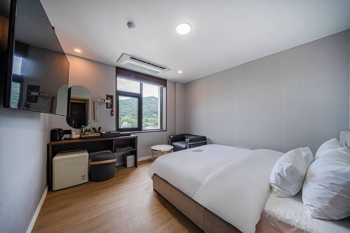 A modern hotel room with a large bed, a desk with a computer, a small table, and a window with a view of greenery.