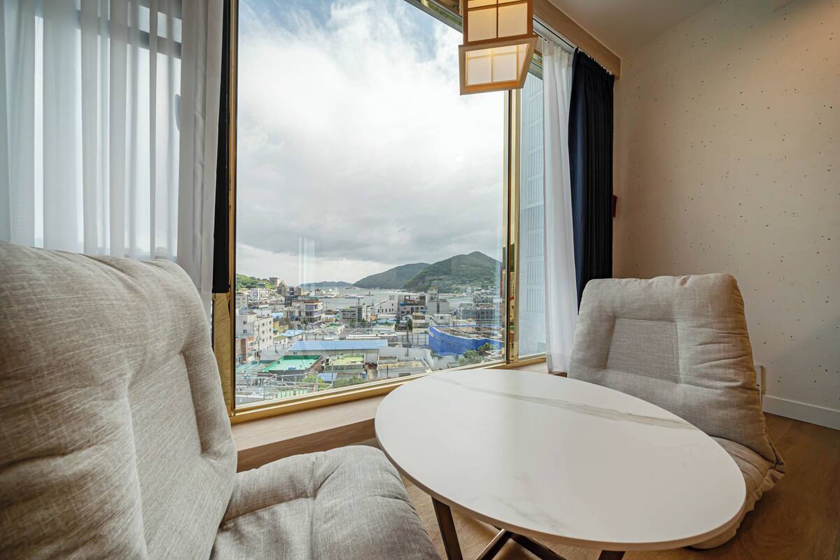 A hotel room with a large window offering a city view, two upholstered chairs, and a small round table.