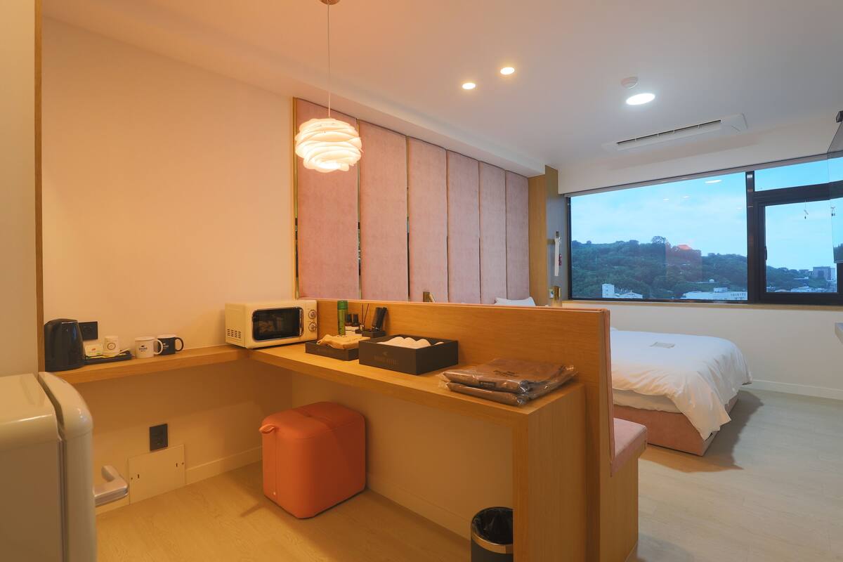 A compact hotel room with a kitchenette, a bed, a window with a city view, and a small seating area.