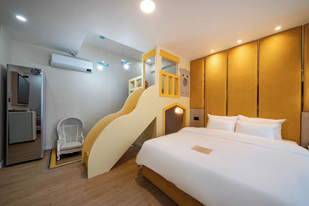 A hotel room with a bed, a desk, a chair, a small desk with a computer, a white chair, a yellow slide, and a wooden wall panel.