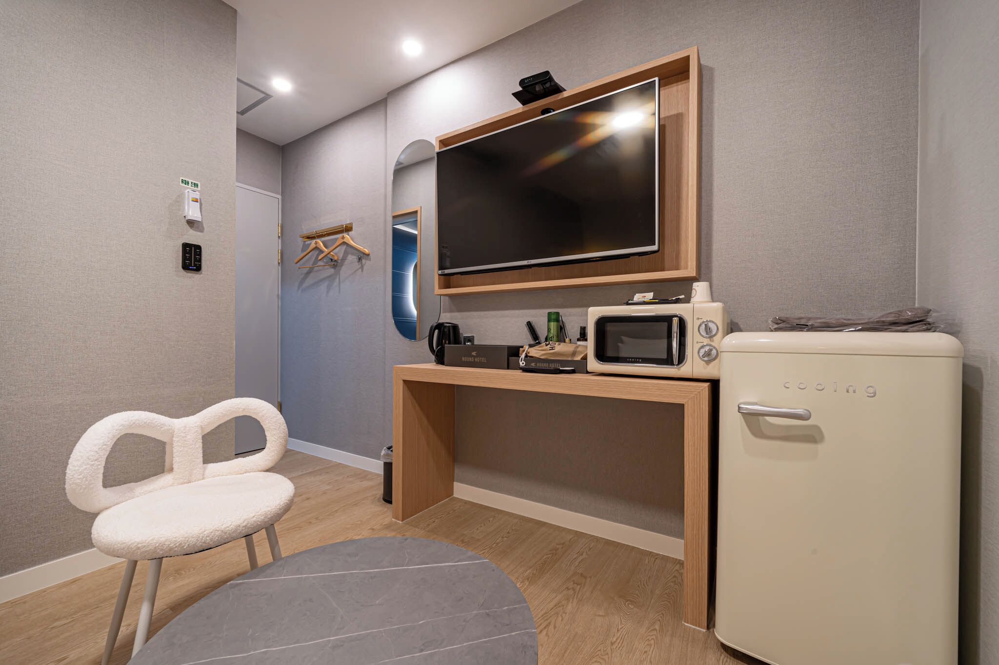A compact room with a flat-screen TV mounted on a wooden shelf, a small refrigerator, a microwave, a chair, and a small table.