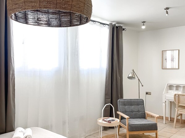 A modern room with a grey armchair, a small side table with a book, a wicker lamp, and a white desk with a chair.