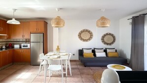 A modern kitchen with wooden cabinets, a stainless steel refrigerator, and a dining area with a white table and chairs. There are two hanging woven light fixtures and decorative woven wall art.