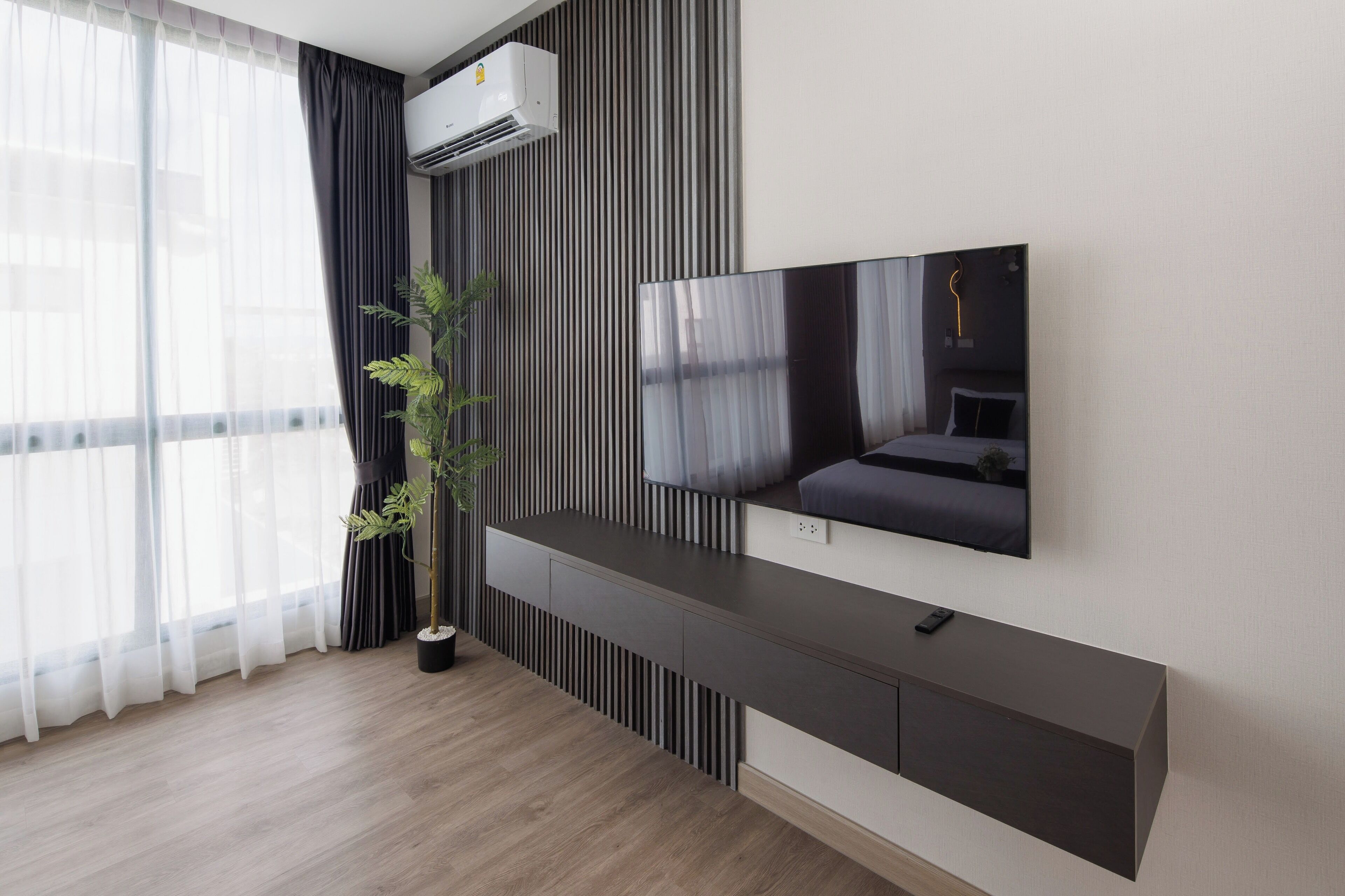 Uma sala de estar moderna com televisão de tela plana na parede, piso de madeira escura, uma janela grande com cortinas leves e um vaso de planta.