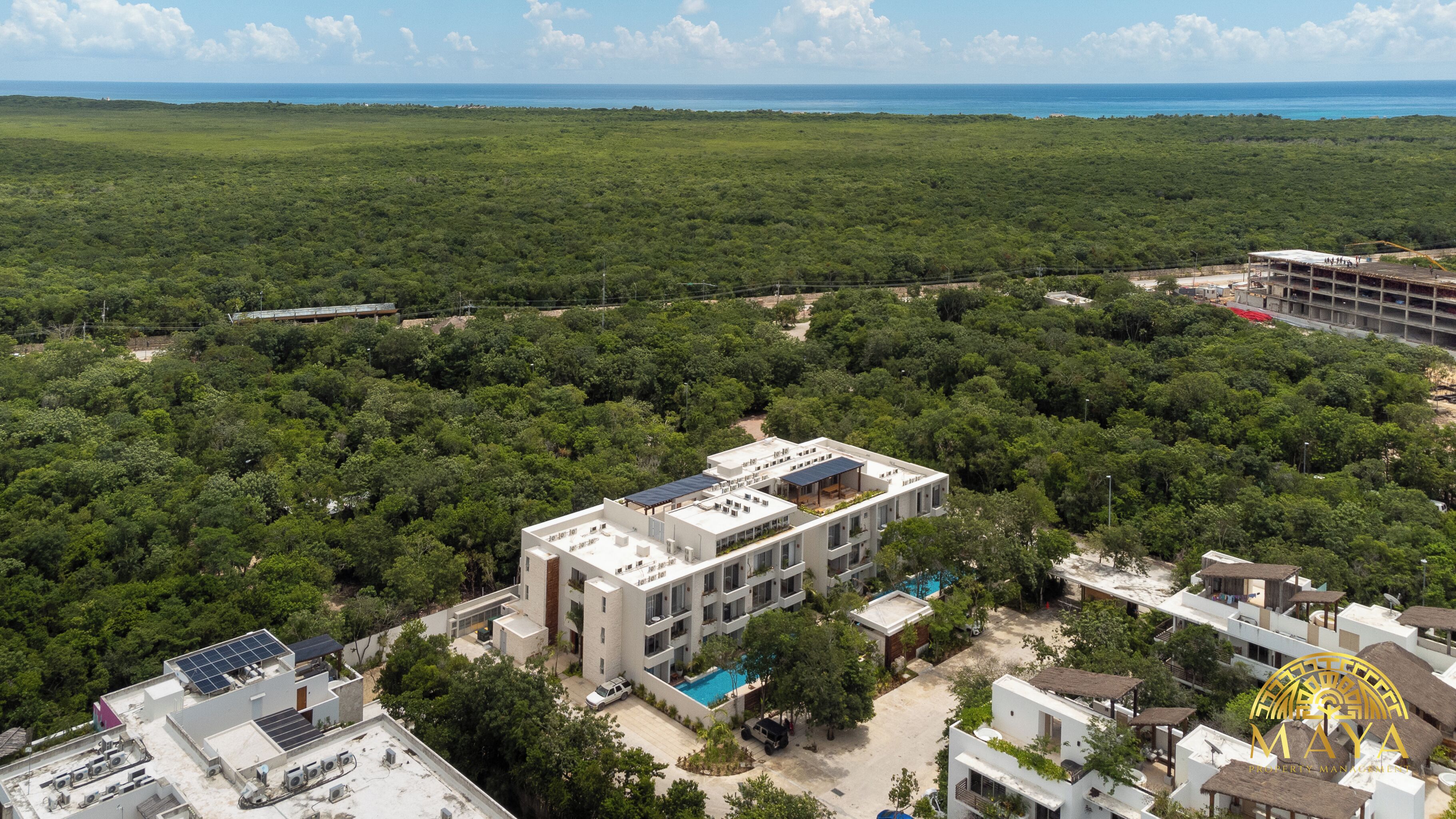 An aerial view of a modern hotel with multiple floors, surrounded by dense greenery and a clear view of the ocean.