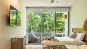 Un dormitorio con una cama, televisión, refrigerador y puerta corrediza de vidrio que da a un balcón con una silla y una planta.
