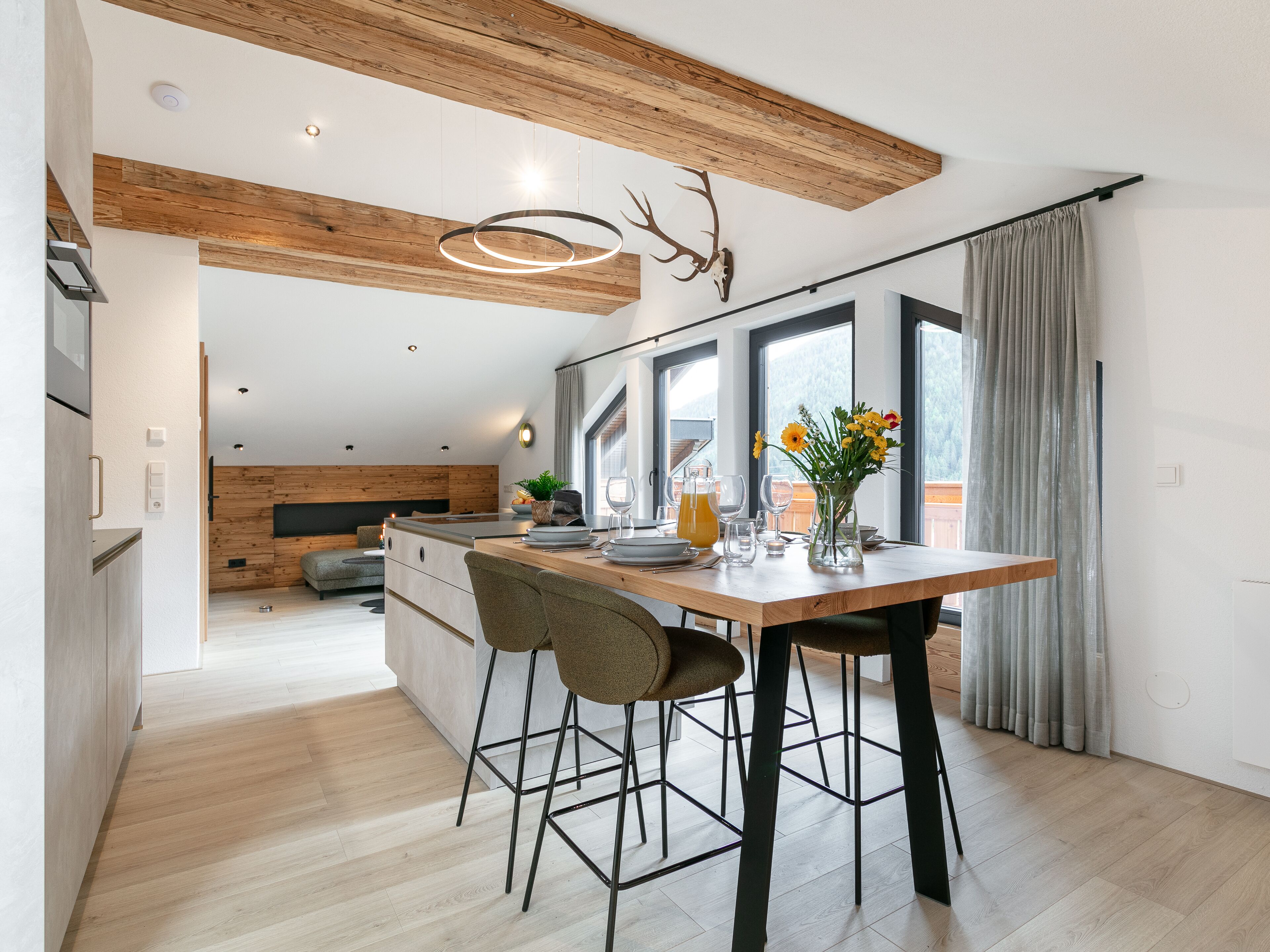Eine moderne Küche mit Holzbalkendecke, einem Essbereich mit zwei Hocker und einem großen Fenster mit Vorhängen.