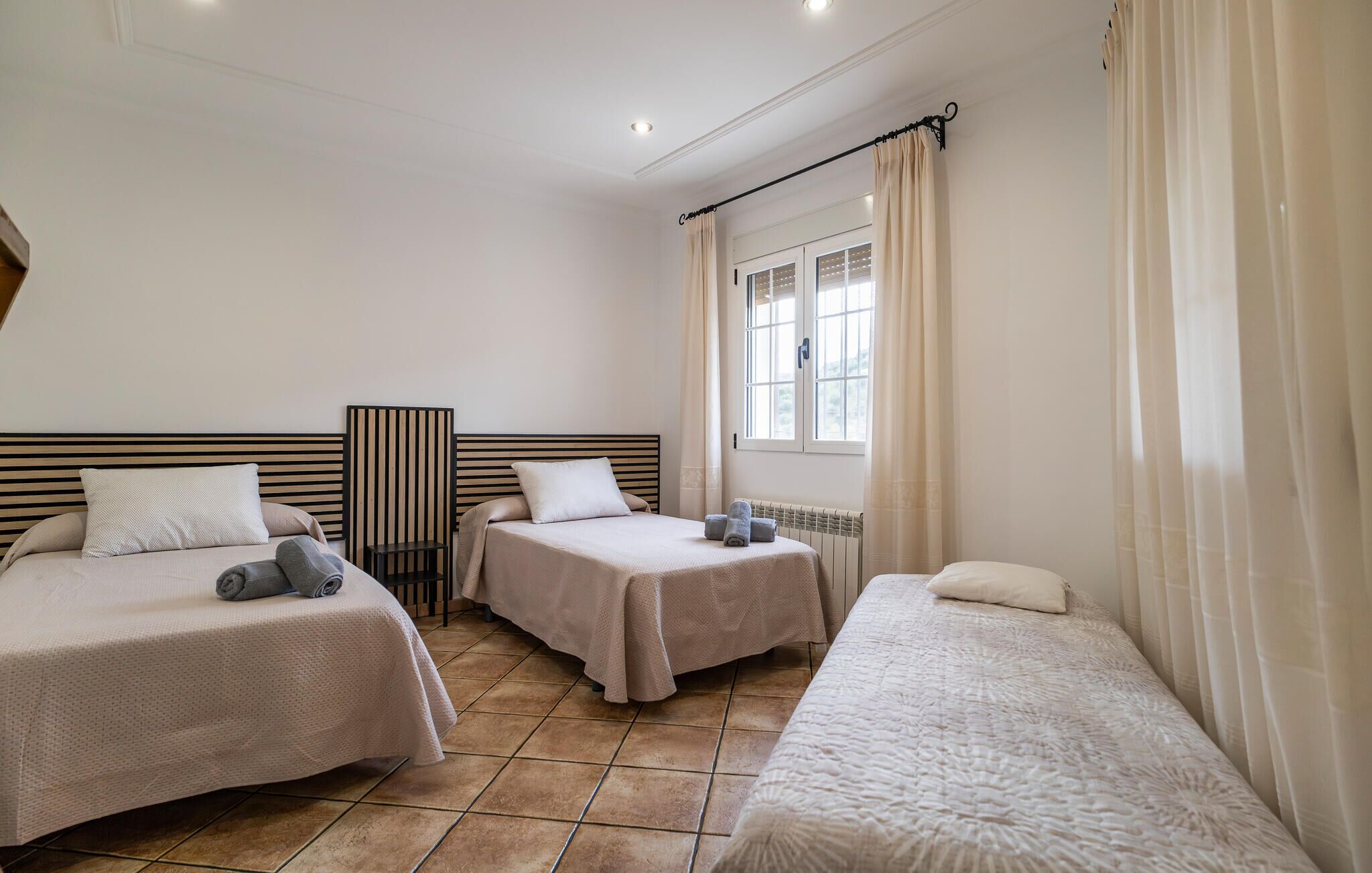 A hotel room with two beds, a window with curtains, a radiator, and a small table with a lamp.