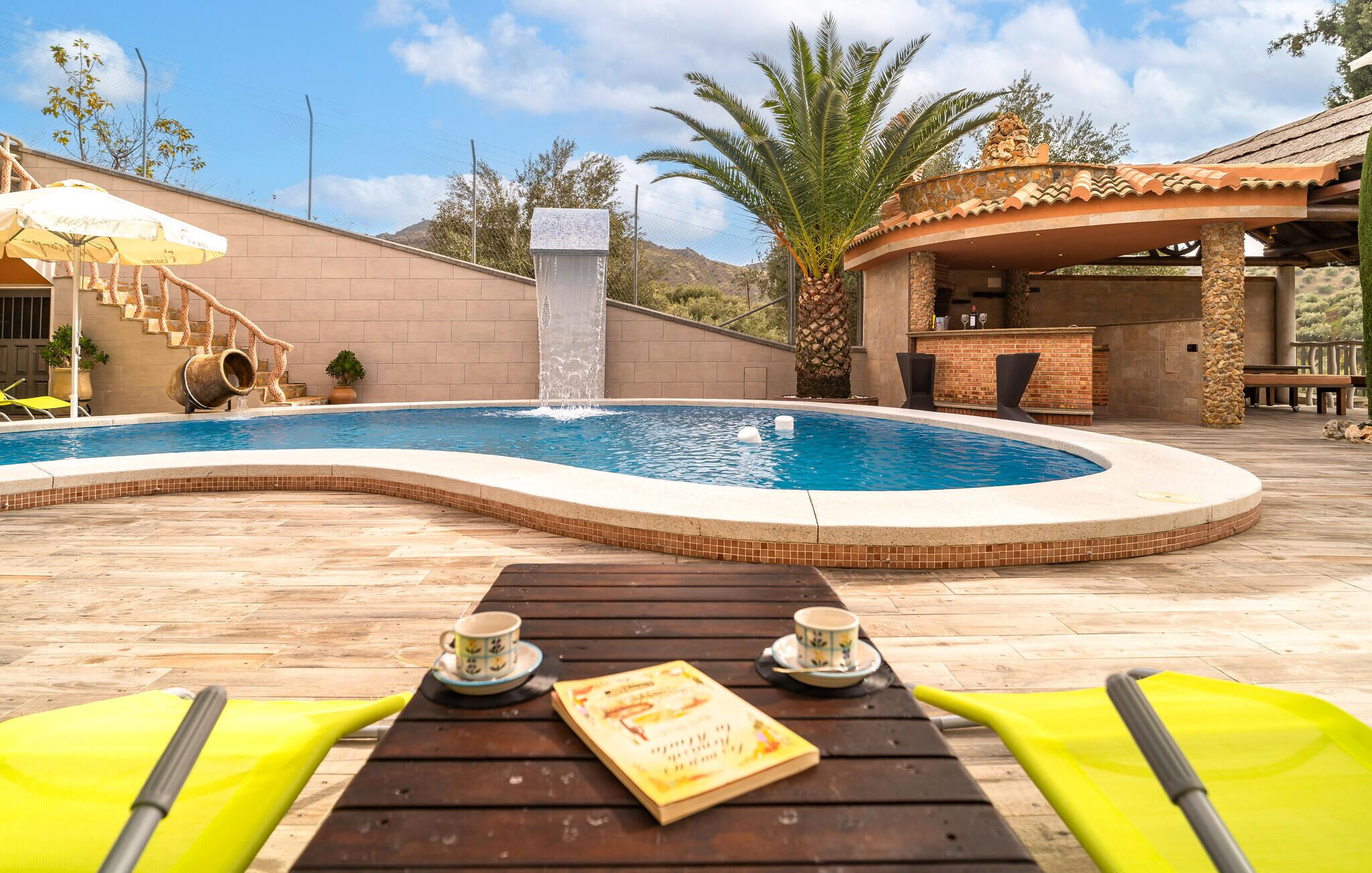 A pool with a waterfall feature, surrounded by a patio area with wooden tables and chairs, and a stone wall.