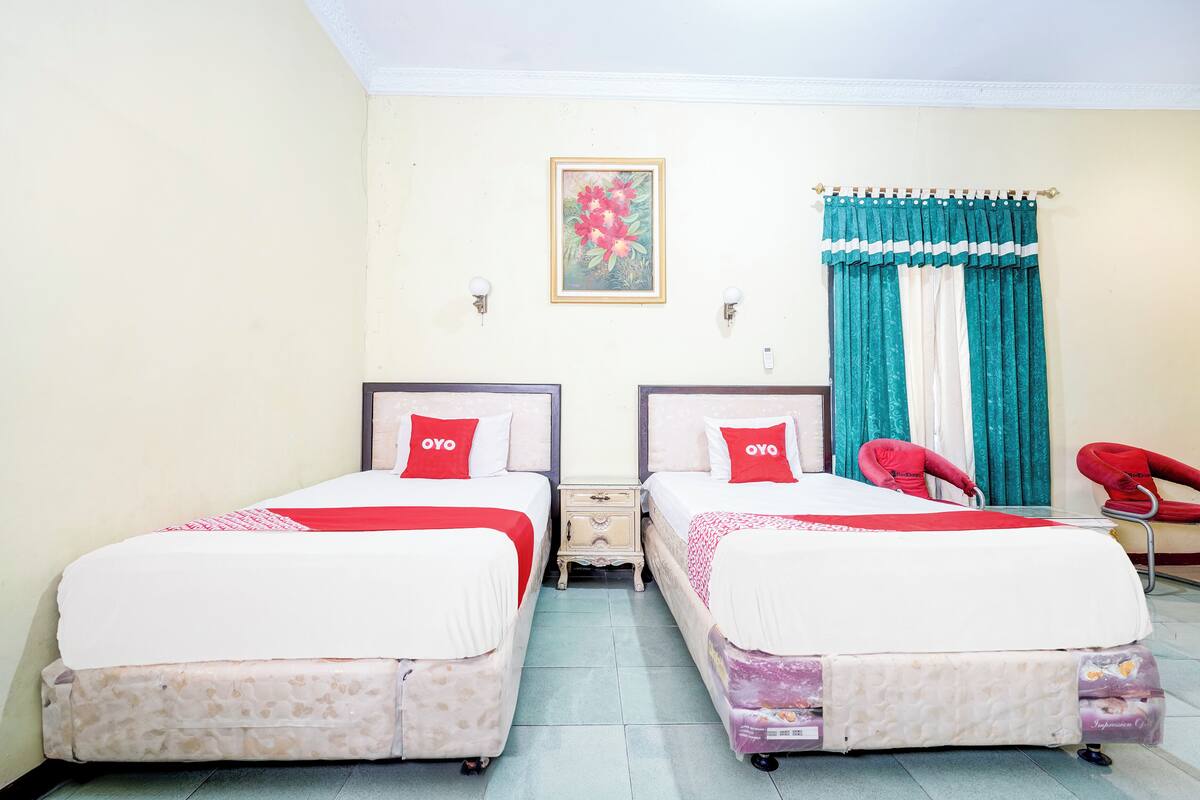A hotel room with two beds, each with a red pillow and a white bedspread. There is a nightstand between the beds, a chair, and a window with teal curtains.