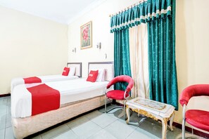 Habitación de hotel con cama doble, dos sillas rojas, una mesita y un ventanal con cortinas.