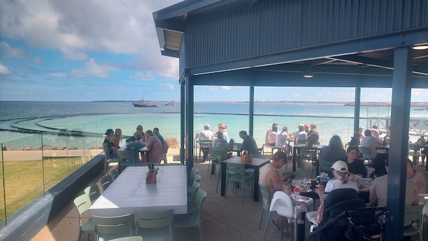 Un espace extérieur aménagé pour les repas, avec des tables et des chaises, offrant une vue sur une étendue d’eau où l’on aperçoit des navires au loin.