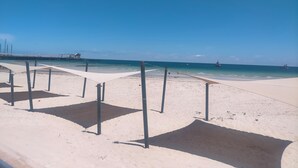 Une plage de sable blanc bordée d’une eau d’un bleu cristallin. On y trouve des poteaux en métal et une promenade en béton.