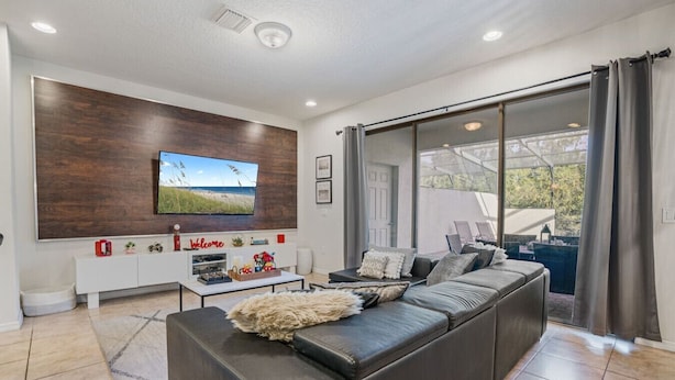 A modern living room with a large sectional sofa, a flat-screen TV mounted on a wooden wall, a white entertainment unit, and a sliding glass door leading to an outdoor area.