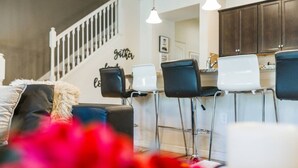 A modern kitchen with bar stools, a staircase, and a 'gather' sign on the wall.