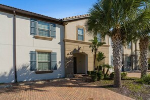 A two-story building with a brick pathway leading to the entrance, surrounded by palm trees and shrubs.