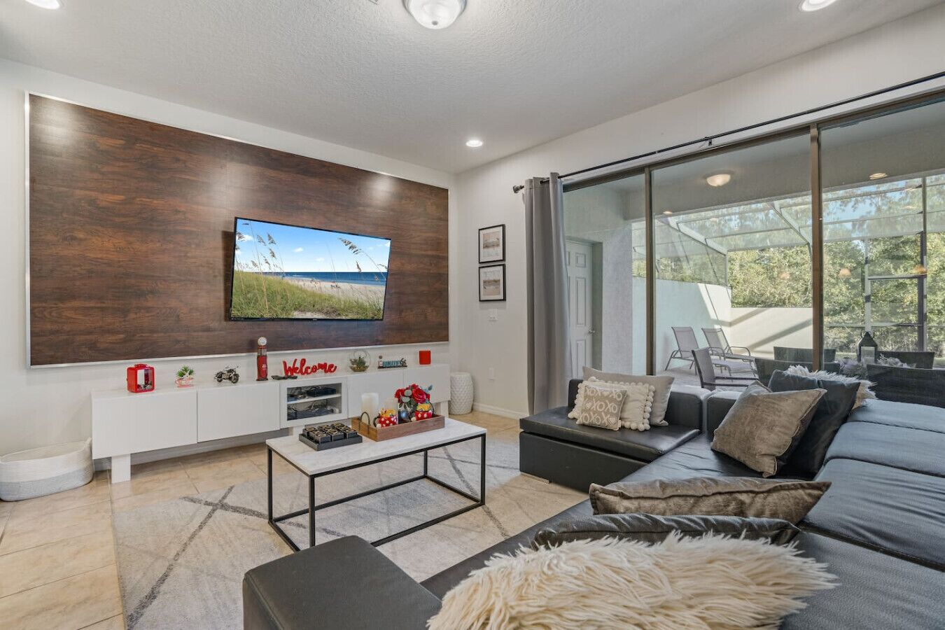 A modern living room with a flat-screen TV, a sofa, a coffee table, and a large window offering a view of the outdoors.