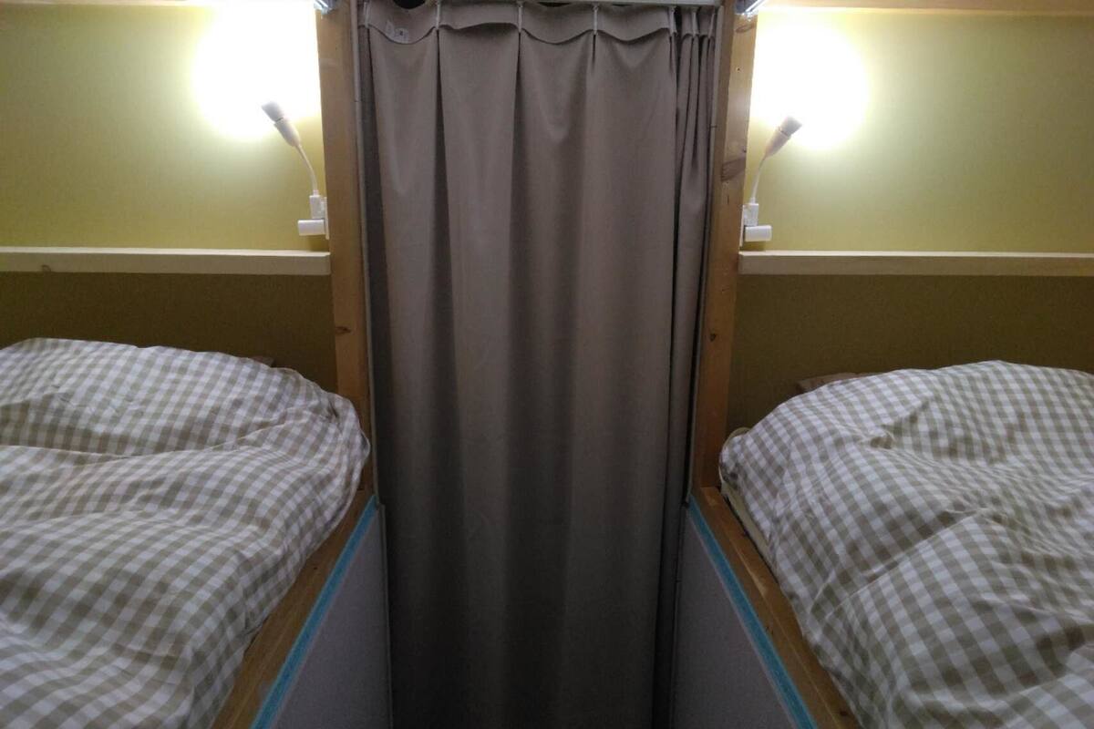 Une chambre avec deux lits simples, chacun recouvert d’une couvre-lit à carreaux, avec un rideau gris et deux lampes de chevet.