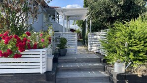 En trägång leder fram till en vit pergola med ett tak av metall, och den är omgiven av frodig grönska och krukväxter.