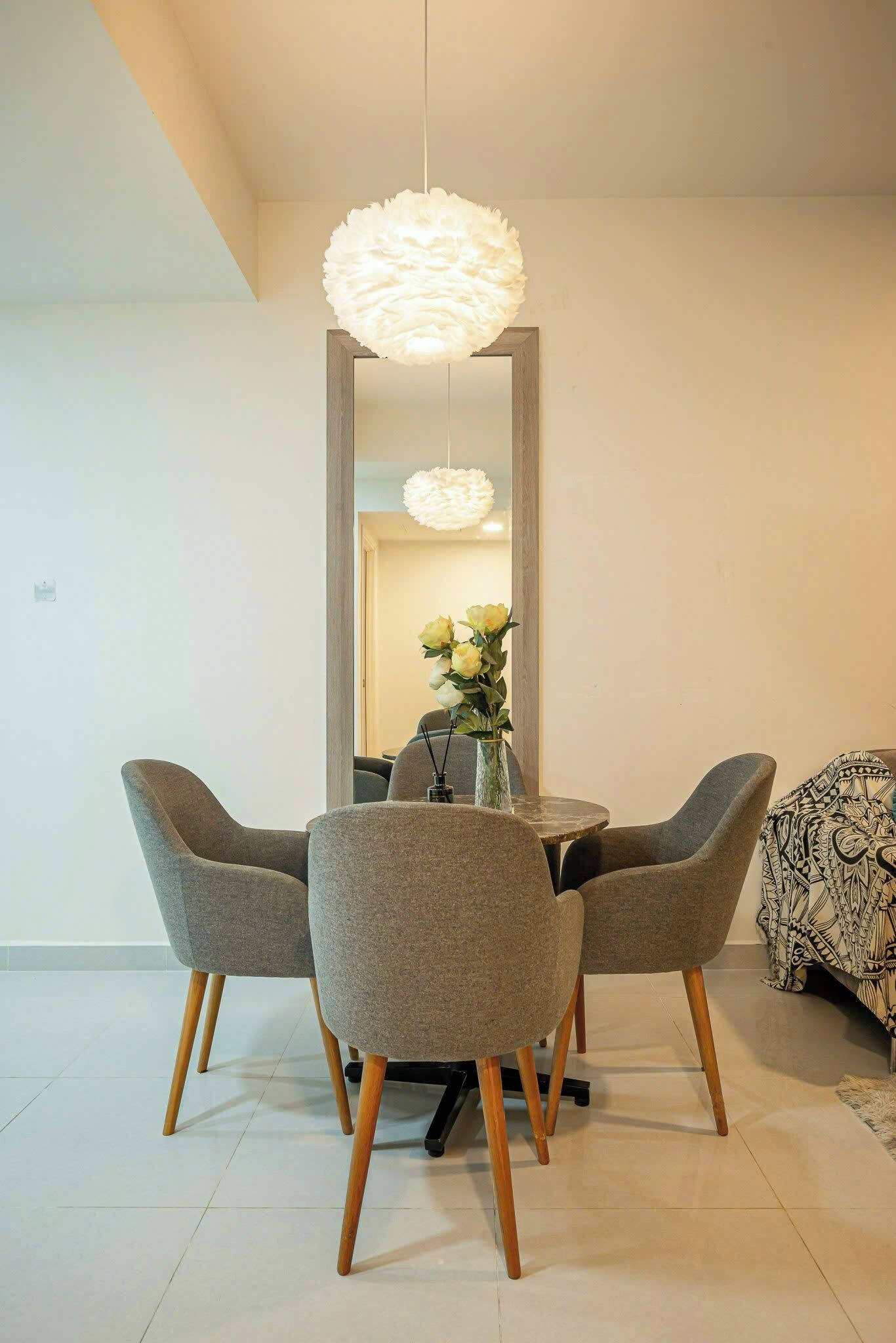 A dining area with a round table, four grey upholstered chairs with wooden legs, a white textured pendant light, and a vase with flowers.