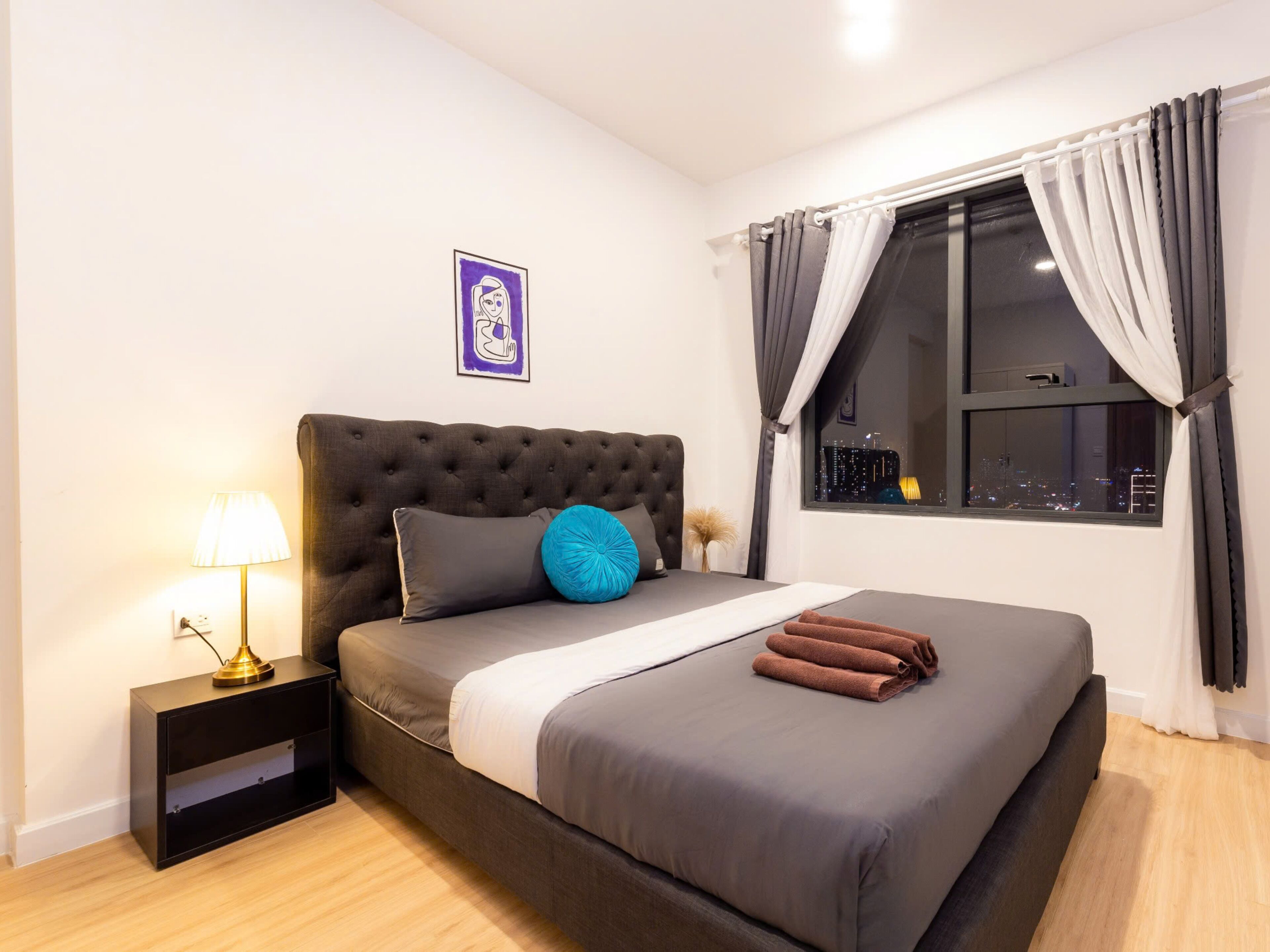 Un dormitorio ordenado con una cama grande, una mesita de noche con lámpara y una ventana con cortinas.