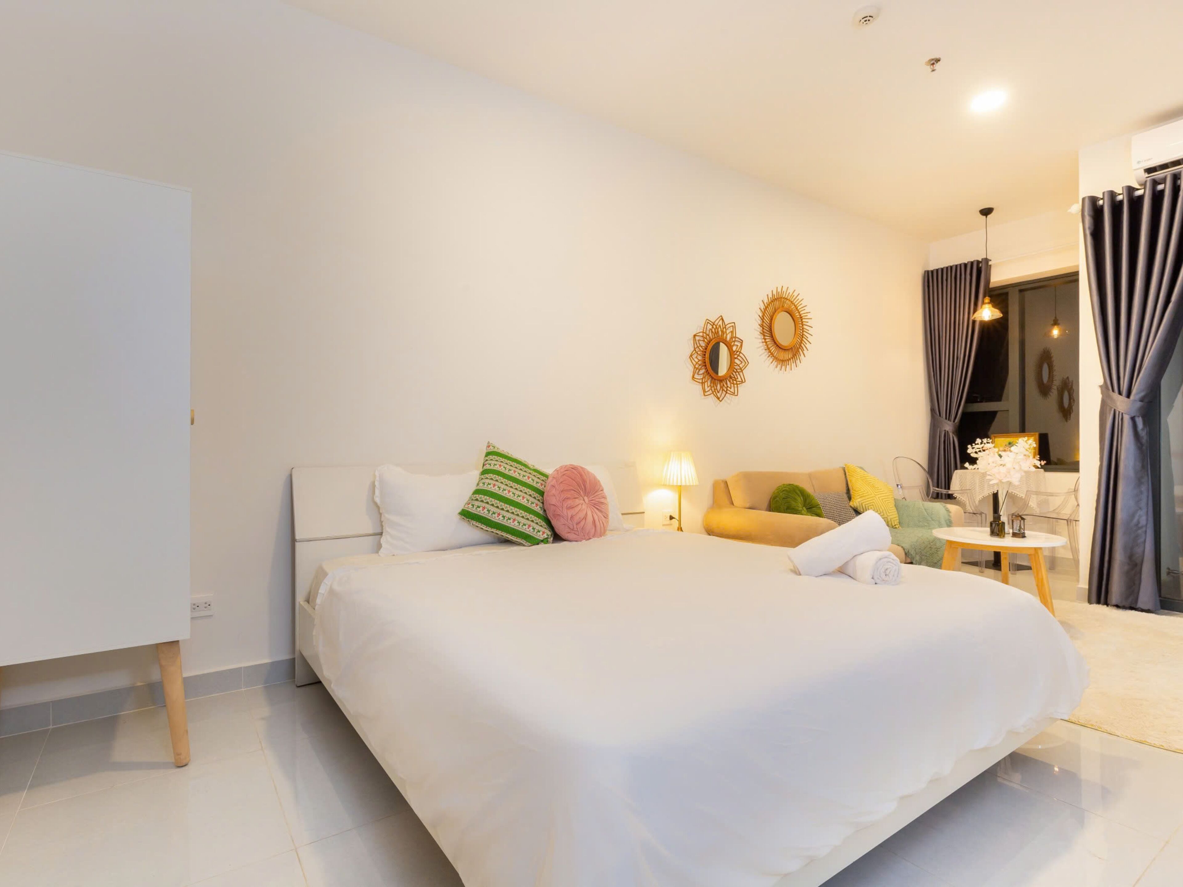 Una habitación de hotel moderna con una cama grande, una mesita de noche con lámpara, un sofá y una pequeña mesa con un jarrón.