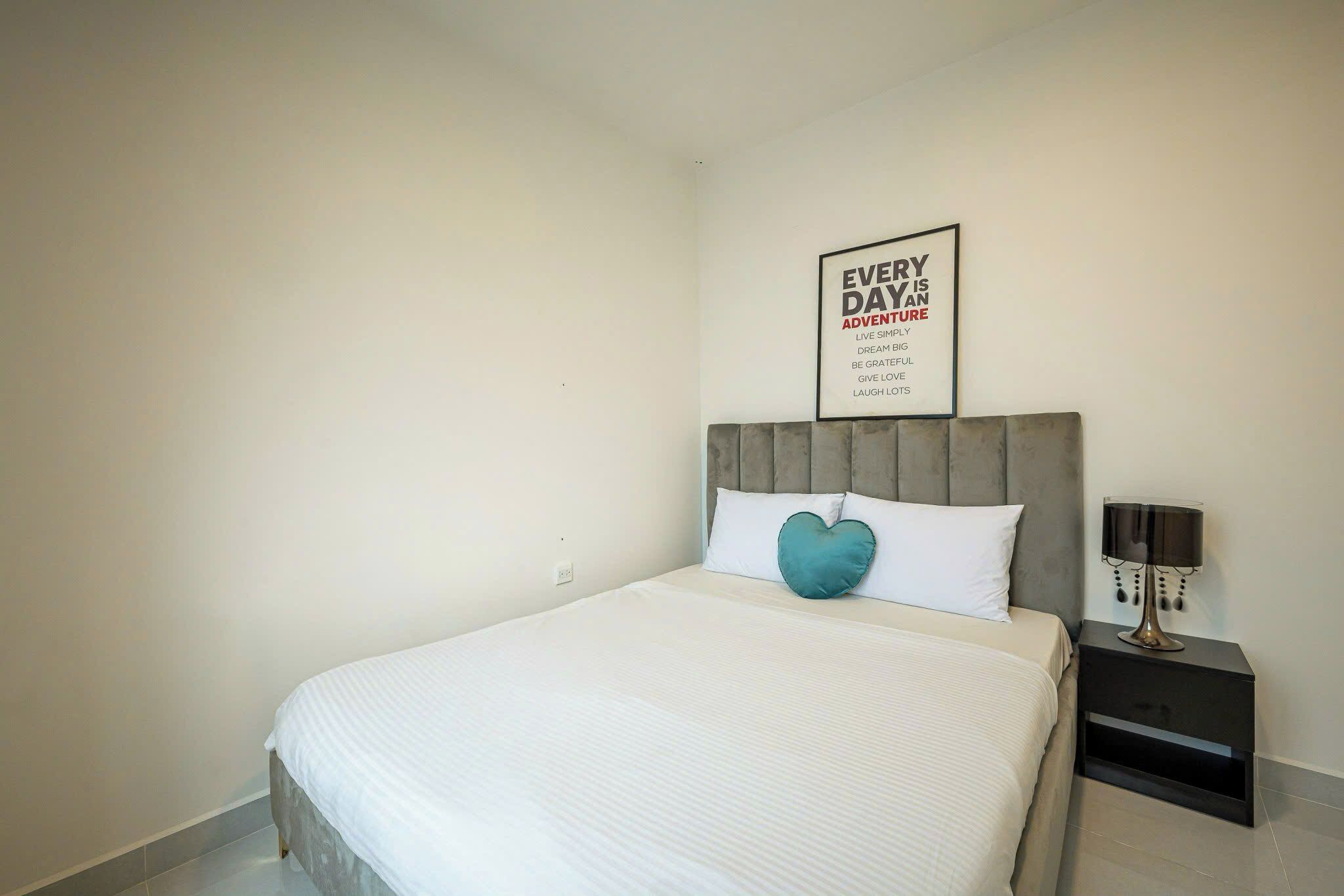 Un lit bien fait, avec une couette blanche et un coussin en forme de cœur de couleur turquoise. Un poster motivant est accroché au mur, au-dessus du lit.