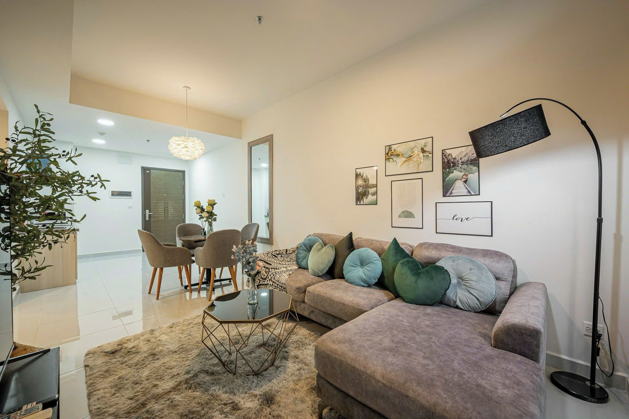 A modern living room with a sofa, coffee table, and decorative pillows. There is a dining area with chairs and a table, and a large plant in the corner.