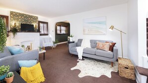 A living room with a grey sofa, a flat-screen TV, a fireplace, and a large painting of a beach. - Renovated 3BR Family Retreat, Sleeps 8 - Dog-Friendly (Akron)
