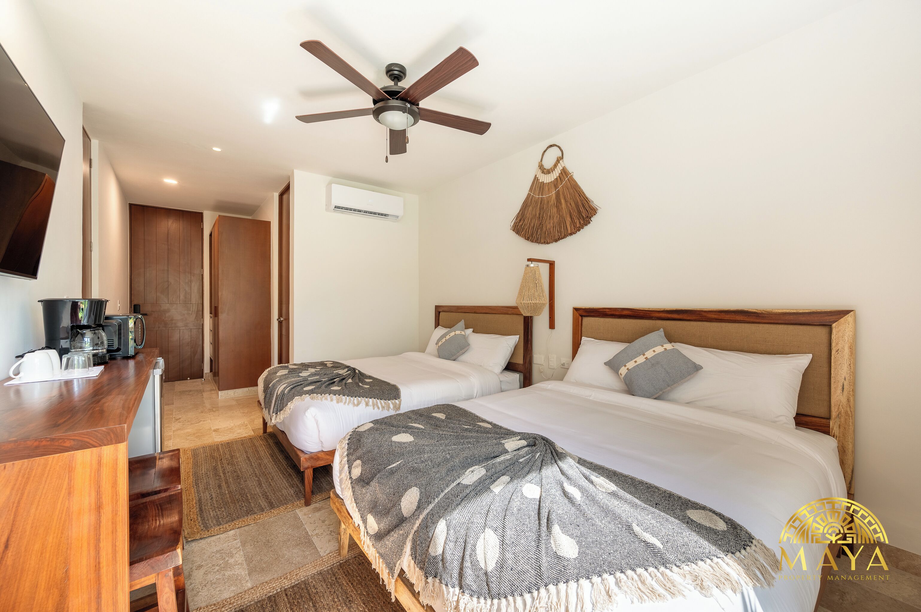 A hotel room with two beds, a ceiling fan, a woven wall hanging, and a kitchenette with a coffee maker.