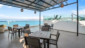 An outdoor patio with a view of the sea, furnished with tables and chairs, and a glass balustrade.