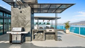 An outdoor patio with a grill and seating area, overlooking a body of water and cityscape.