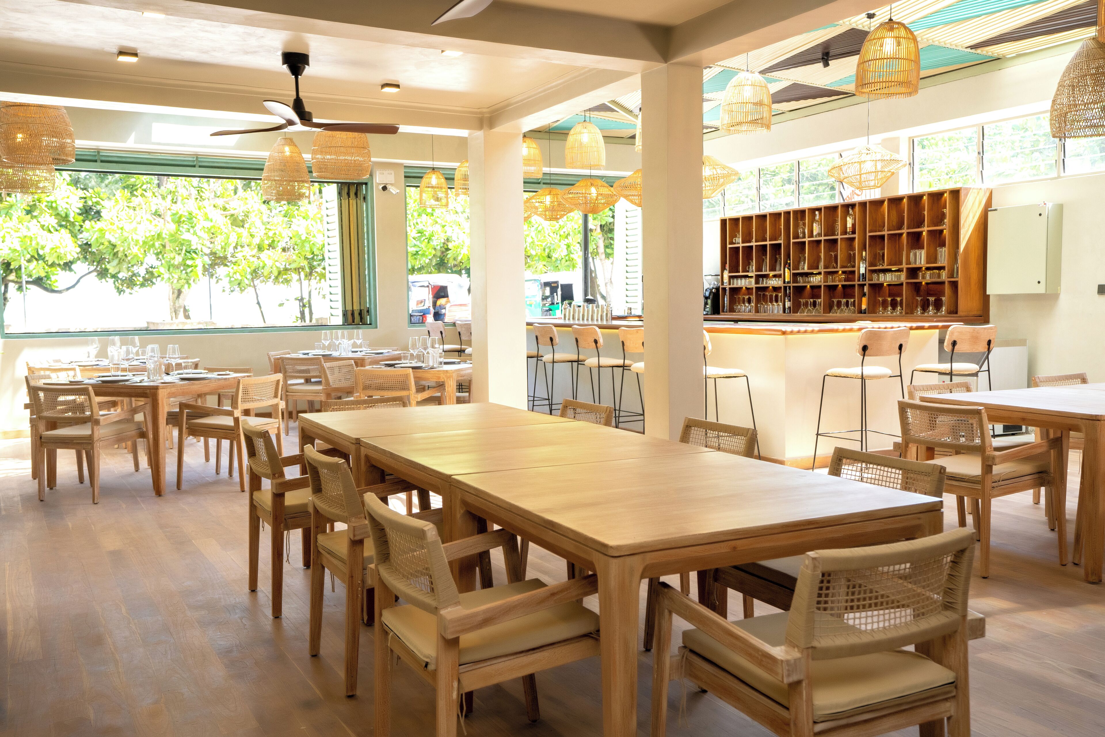 Un restaurant spacieux avec des tables et des chaises en bois, de grandes fenêtres donnant sur la verdure et un bar avec des étagères remplies de bouteilles.